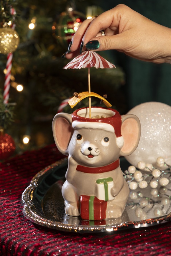 A grey mouse mug in a red Santa hat and scarf, holding a present, with a candy-stripe umbrella.
