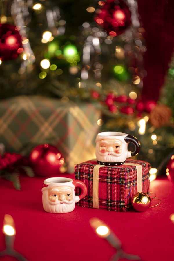 Two small Santa-face shot mugs, one winking with a black handle and one smiling with a red handle, beside a tartan-wrapped present.