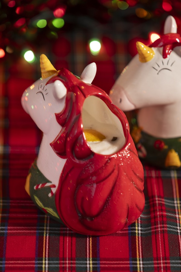 Two white unicorn mugs with flowing red manes, gold horns and ornament-printed bases, on a tartan cloth under Christmas lights.
