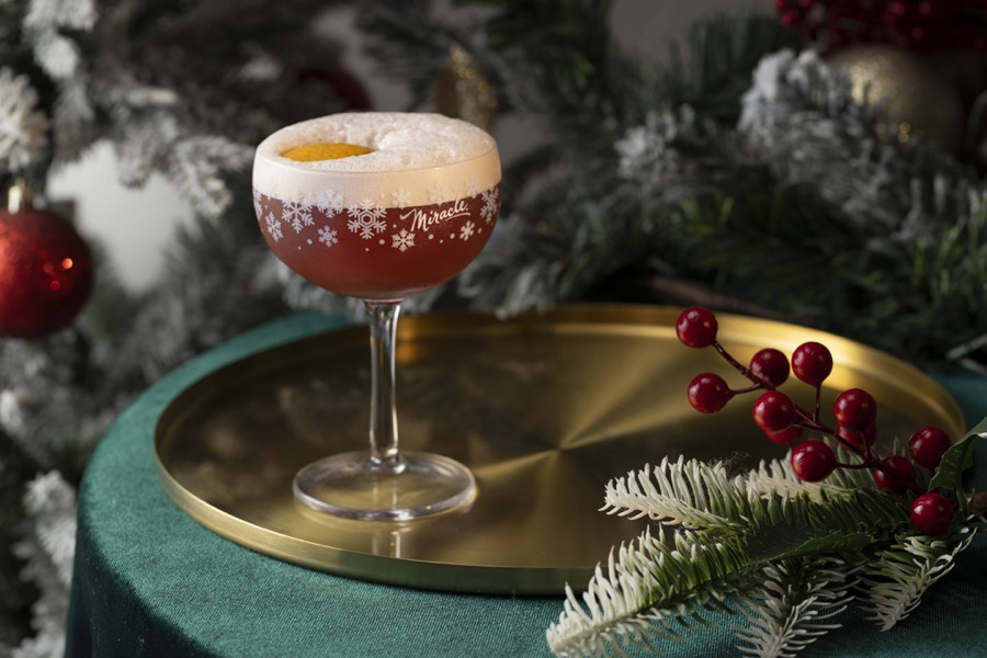 A red foamed cocktail in a snowflake-printed Miracle coupe on a gold tray, beside frosted pine and berries.