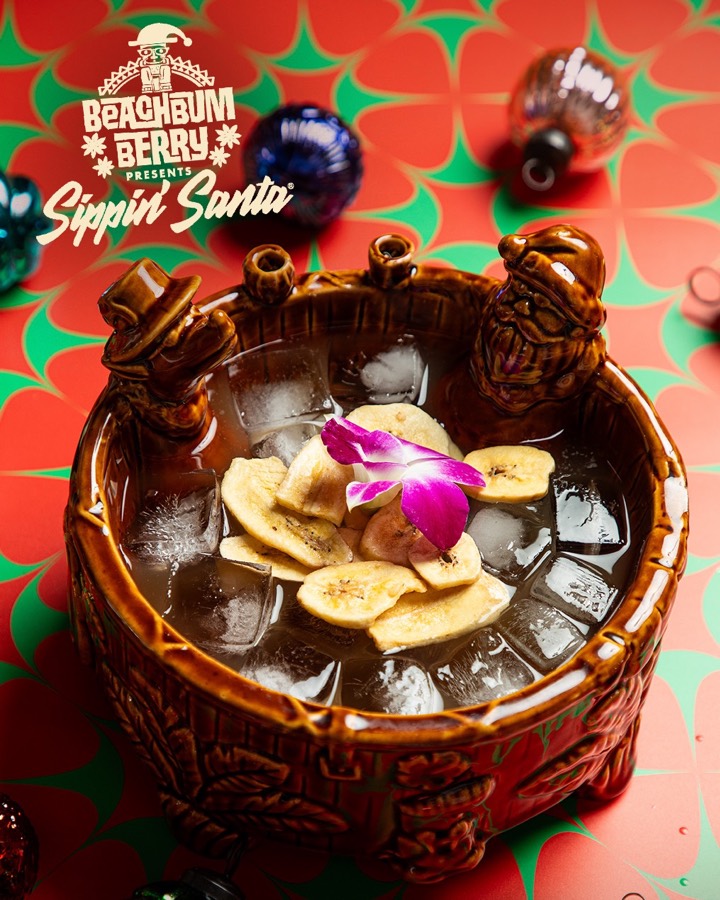 A large brown tiki punch bowl topped with banana chips, ice and an orchid, on a red and green background.