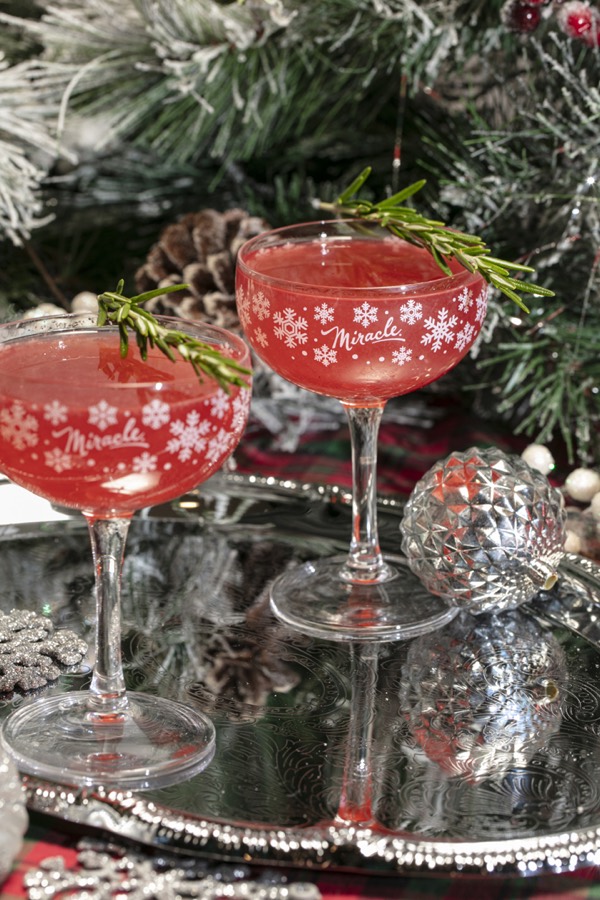 Two Christmapolitans in snowflake-printed Miracle coupes with rosemary sprigs, on a silver tray beside a frosted tree.